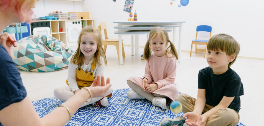 Three children sit cross-legged on a playmat in a classroom as an adult tosses a ball to a child sitting with his hands cupped