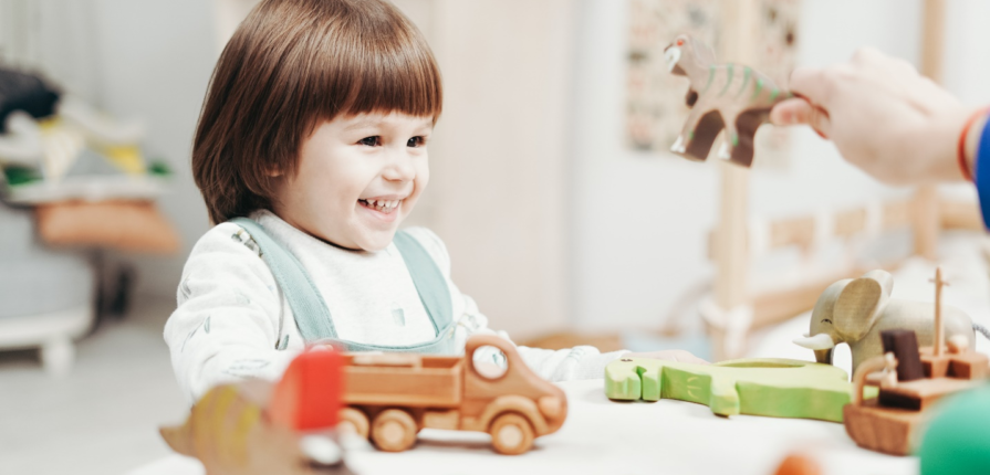 A toddler smiles wide while playing with wooden toys on a table as an adult hand comes in from the right side holding a wooden dinosaur