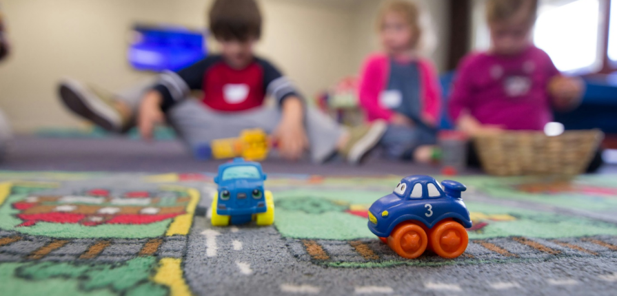 Two vibrant toy cars on a road-themed carpet, with blurred preschoolers in the background at a day nursery.