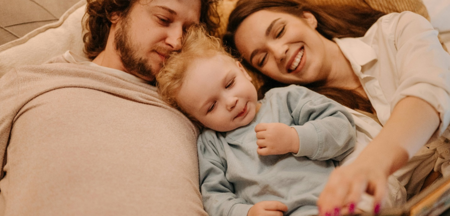 Bedtime stories for young children are great for bonding, as can be seen here with happy parents lying with a toddler between them in bed as they read a story.