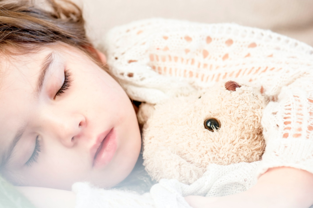 A toddler in a crocheted top peacefully asleep, clutching a plush light brown teddy bear, highlighting the significance of mastering toddler sleep for a cozy bedtime routine.