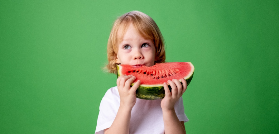 A charming toddler in a white shirt enjoying a slice of watermelon against a vibrant green backdrop, promoting a healthy and refreshing addition to the toddler diet.