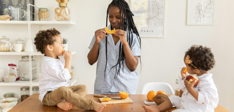 An infant and toddler sit on a table eating sliced oranges as their mother stands beside them, having learned how to encourage healthy eating through a toddler nutrition guide.