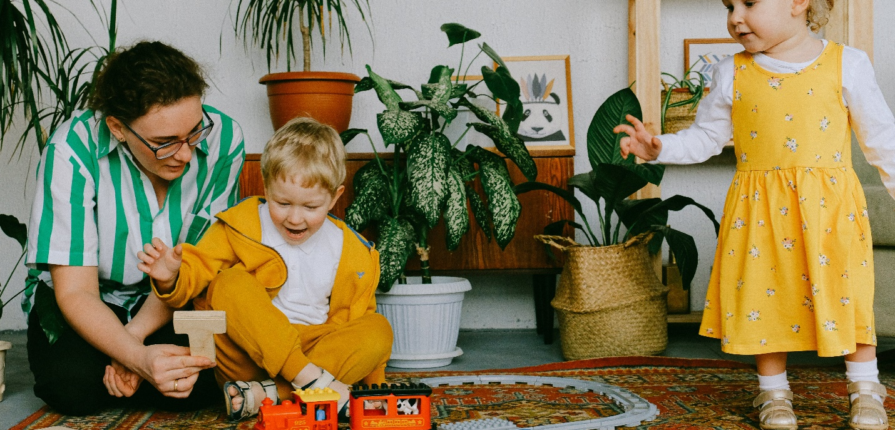 A toddler in a yellow dress stands beside another seated toddler and their mother, engaged in playful exploration with a train and track set amid lush plants, showcasing how the right infant and toddler toys aid in development.