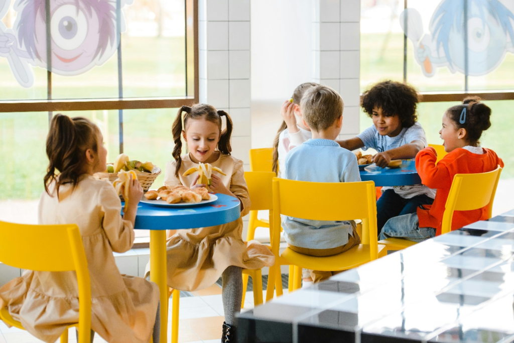 Four children sit around a blue table on yellow chairs eating together, with two girls sitting at another blue table eating bananas at a school cafeteria, stressing the need for toddler nutrition guides in daycares and schools.