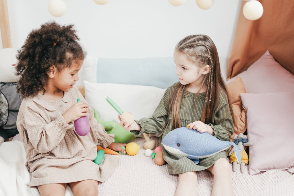 Two preschool-aged girls joyfully share wooden vegetable toys on a plush surface, illustrating positive interactions in disciplining a toddler's developing behavior.