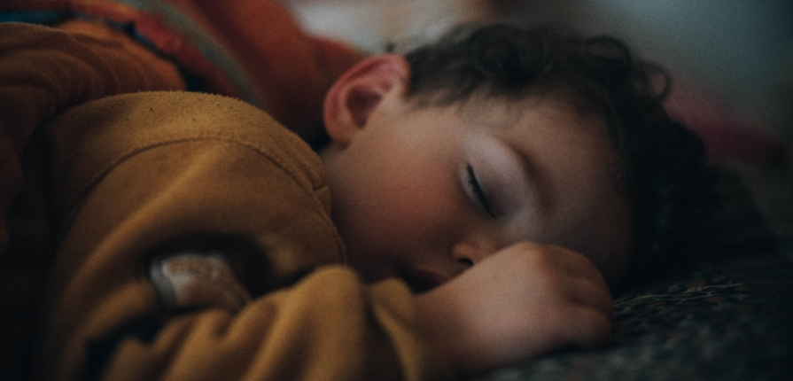 A peaceful toddler, dressed in an ochre top, sleeping soundly on his stomach under a cozy blanket, showcasing the importance of a well-rested toddler for overall health, shedding light on the importance of mastering toddler sleep.