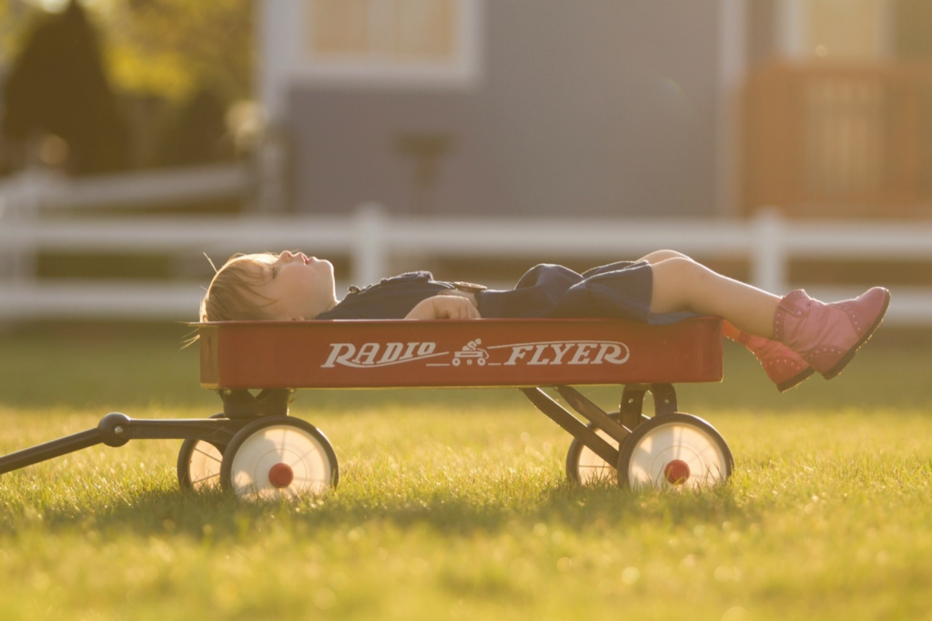 A sleeping toddler in a blue dress and pink boots peacefully napping in a red Radio Flyer wagon amidst the grass, showcasing natural toddler sleep patterns.