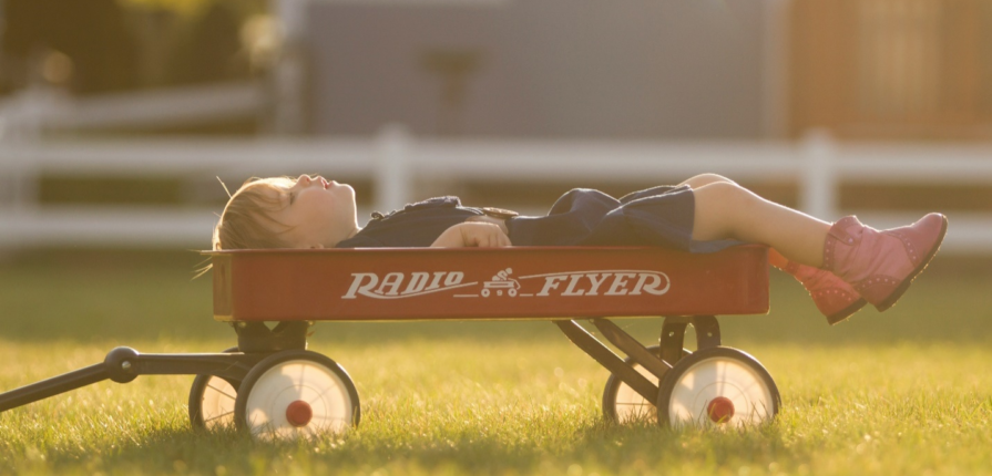 A sleeping toddler in a blue dress and pink boots peacefully napping in a red Radio Flyer wagon amidst the grass, showcasing natural toddler sleep patterns.