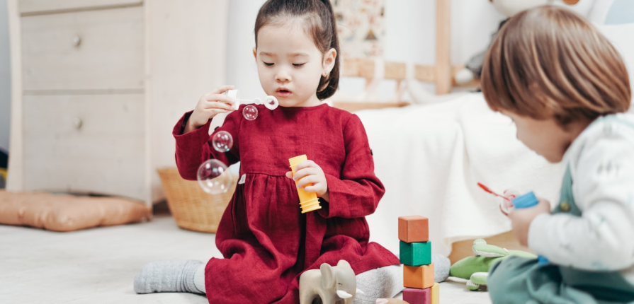 Toddler in a red burgundy dress sits on the floor blowing bubbles while another toddler explores building blocks, fostering social skills and emotional intelligence - building EQ in toddlers.