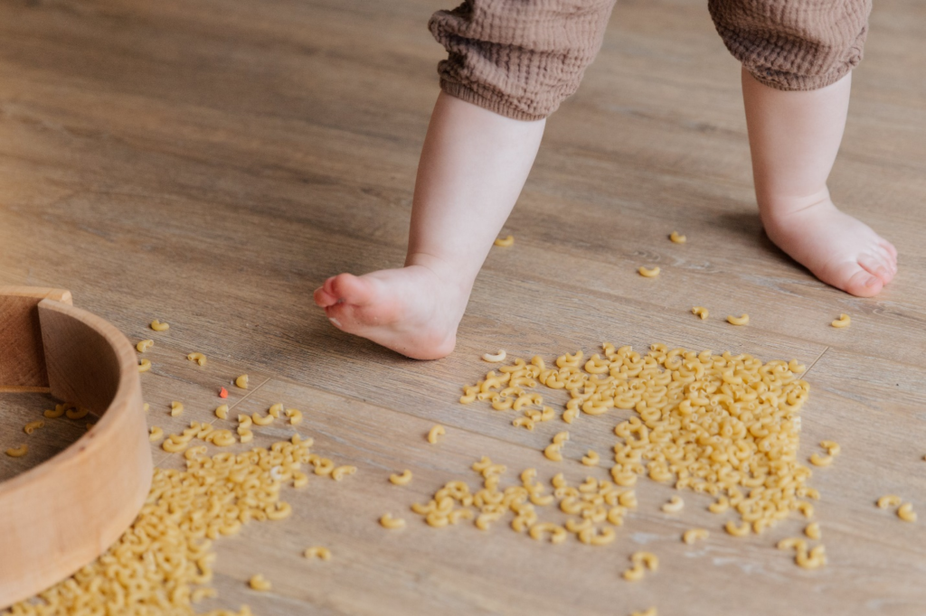 Close-up of toddler's feet on a floor scattered with macaroni - a delightful mess of exploration and play, making for an engaging activity for toddlers.