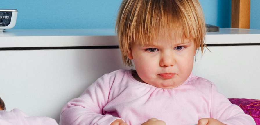 A toddler with blonde hair in a pink t-shirt looking grumpy as another child's hand extends onto her blanket, showcasing what anger in children can look like.