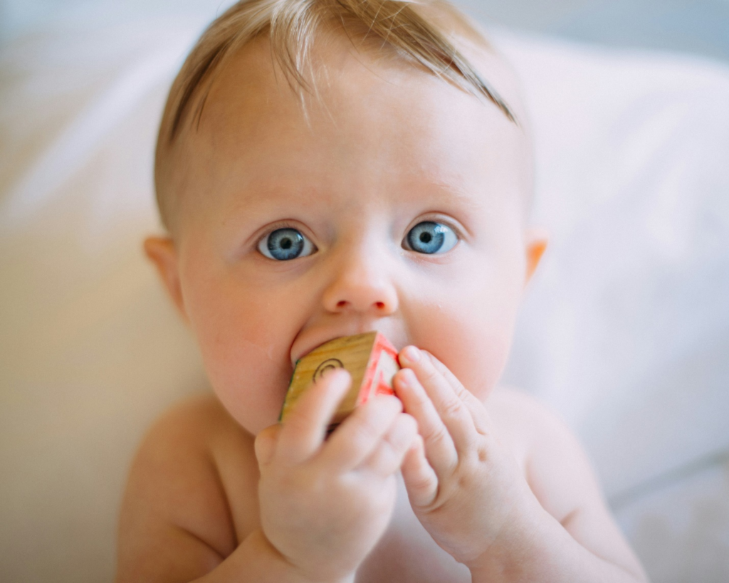 An infant with bright blue eyes and light hair putting a wooden cube in his mouth