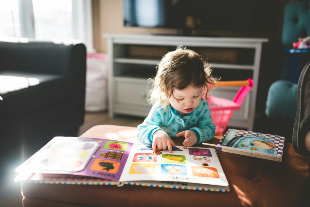 A toddler with a curious gaze looks at a book on a table in a sunlit living room, nurturing toddler self-esteem with early exploration.