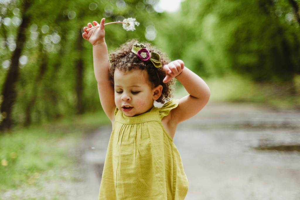 A toddler in a green dress joyfully holding up a dandelion outdoors, illustrating how positive parenting tips can do wonders for a child’s confidence.
