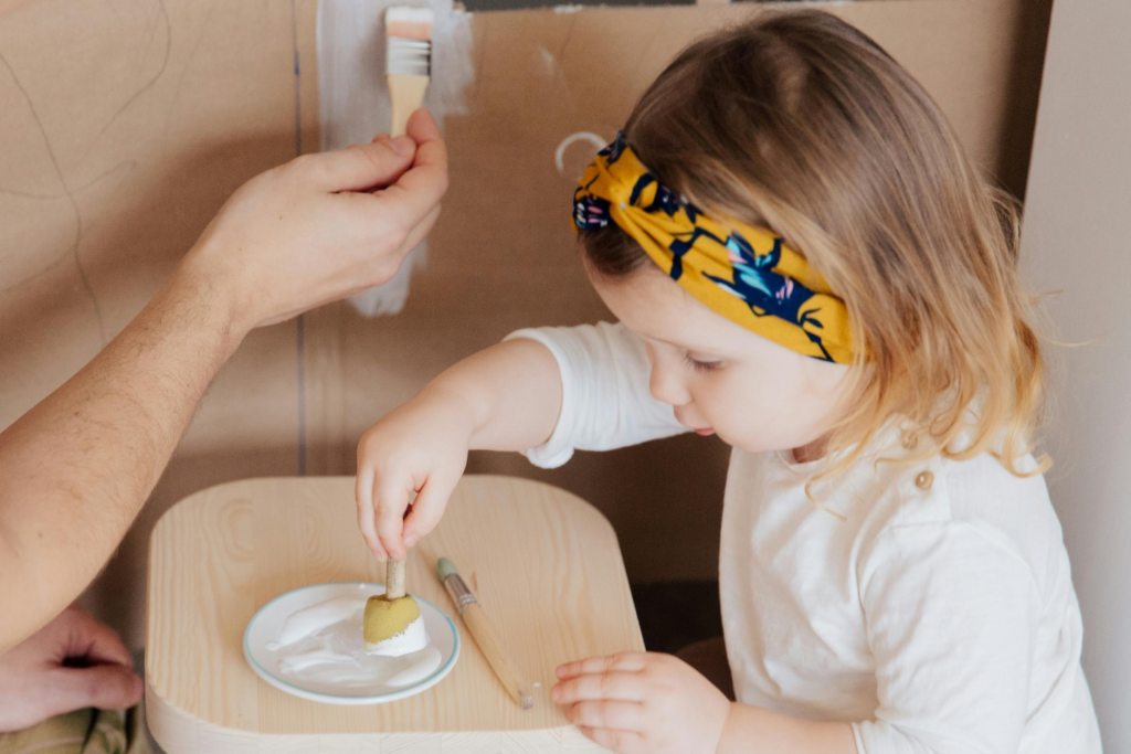 A little girl in white, wearing a yellow floral headband, dips a foam stamp in white paint on a saucer while an adult hand paints cardboard with a brush, showing that sensory play for toddlers can get messy but is also a great way to bond.
