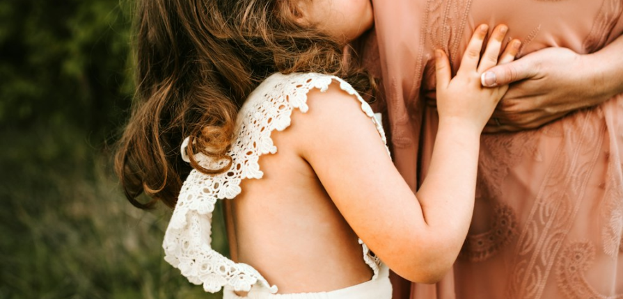 A little girl in a white dress kissing her mom’s pregnant belly
